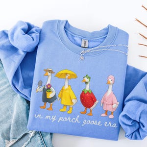 May include: A light blue sweatshirt with a whimsical graphic of four geese dressed in various outfits. The geese are wearing a hat, raincoat, strawberry, and robe. The text "in my porch goose era" is printed below the geese.