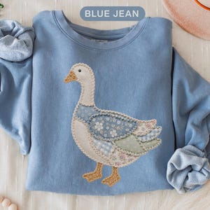 May include: A blue jean colored crewneck sweatshirt featuring a large applique of a goose. The goose is constructed from various fabric patches in floral, gingham, and solid patterns with visible stitching. The text BLUE JEAN is printed above.