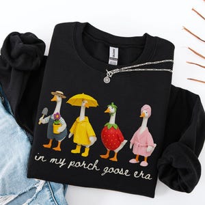 May include: Black sweatshirt featuring a whimsical design of four geese dressed in various outfits. One goose holds a trowel and flower pot, another wears a yellow raincoat, a third is a strawberry, and the last wears a pink robe. The text "in my porch goose era" is below.