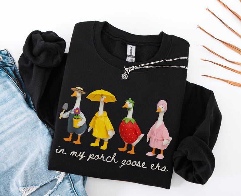 May include: Black sweatshirt featuring a whimsical design of four geese dressed in various outfits. One goose holds a trowel and flower pot, another wears a yellow raincoat, a third is a strawberry, and the last wears a pink robe. The text "in my porch goose era" is below.