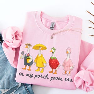 May include: A light pink sweatshirt featuring a whimsical design of four geese dressed in various outfits. One goose holds a trowel and flower pot, another wears a yellow raincoat and umbrella, and the others are dressed as a strawberry and in a pink robe. The text "in my porch goose era" is printed below.