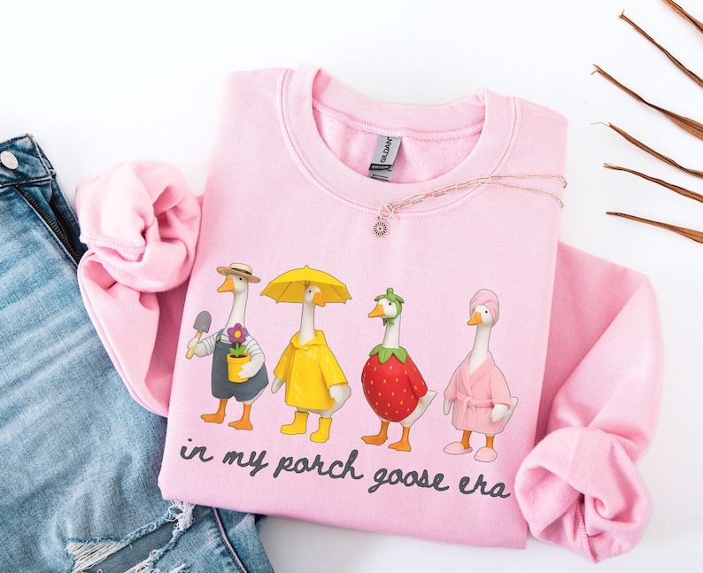 May include: A light pink sweatshirt featuring a whimsical design of four geese dressed in various outfits. One goose holds a trowel and flower pot, another wears a yellow raincoat and umbrella, and the others are dressed as a strawberry and in a pink robe. The text "in my porch goose era" is printed below.
