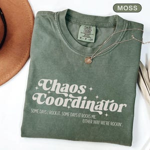 May include: A moss green t-shirt with the words "Chaos Coordinator" in a retro font. The shirt also has the phrases "Some days I rock it. Some days it rocks me. Either way we're rockin'." A gold necklace with a pendant rests on the shirt.
