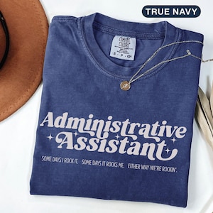 May include: A navy blue t-shirt with the words "Administrative Assistant" in a retro font. The shirt also has the text "SOME DAYS I ROCK IT. SOME DAYS IT ROCKS ME. EITHER WAY WE'RE ROCKIN'". A gold necklace with a pendant is on the shirt.