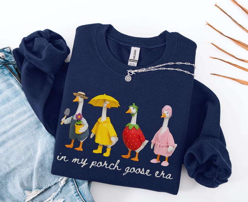 May include: Navy blue sweatshirt featuring a whimsical illustration of four geese dressed in various outfits. One goose holds a trowel and flower pot, another wears a yellow raincoat, a third is in a strawberry costume, and the last wears a pink robe. The text "in my porch goose era" is below the illustration.