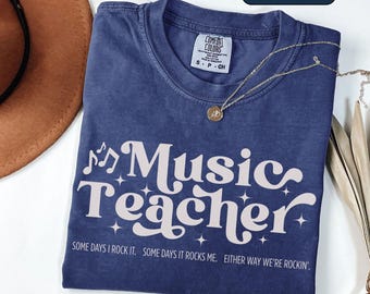 May include: True navy blue t-shirt with the text "Music Teacher" in a retro font, accompanied by musical notes and stars. The shirt also includes the phrases "Some days I rock it. Some days it rocks me. Either way we're rockin'." A gold necklace rests on the shirt.