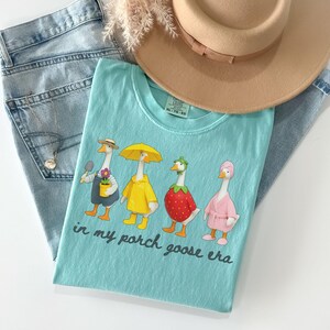 Pu&ograve; includere: Una maglietta azzurra con una grafica di quattro oche in vari outfit, tra cui un giardiniere, un impermeabile, una fragola e un accappatoio. Il testo "in my porch goose era" &egrave; stampato sotto. La maglietta &egrave; abbinata a jeans blu chiaro e un cappello beige.