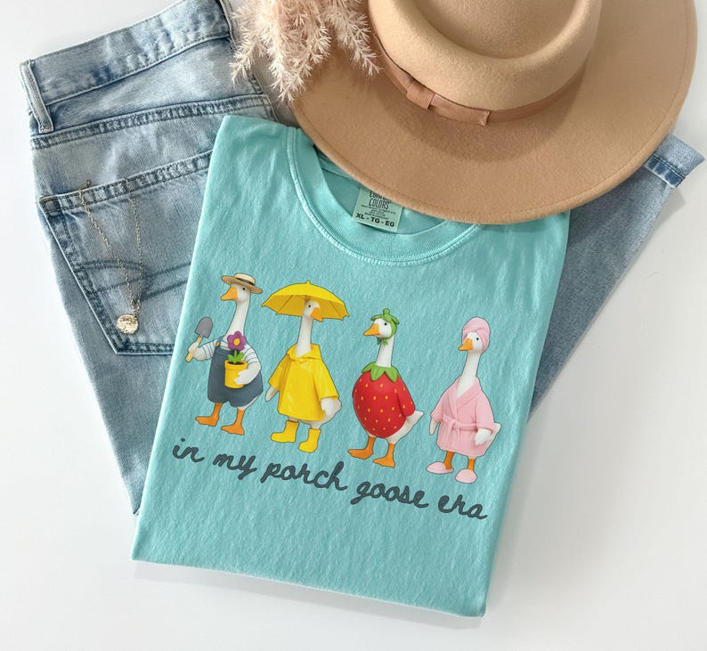 Pu&ograve; includere: Una maglietta azzurra con una grafica di quattro oche in vari outfit, tra cui un giardiniere, un impermeabile, una fragola e un accappatoio. Il testo "in my porch goose era" &egrave; stampato sotto. La maglietta &egrave; abbinata a jeans blu chiaro e un cappello beige.
