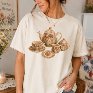 May include: A cream-colored short-sleeve t-shirt featuring a graphic print of a patchwork-style teapot, a teacup on a saucer, a creamer, a sugar bowl, and a small heart-shaped cookie on a plate, all decorated with brown and tan floral patterns.