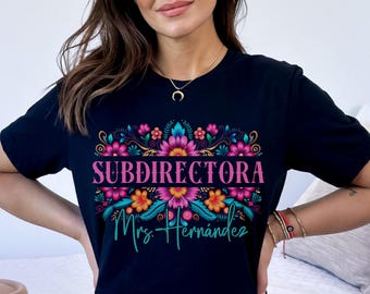 May include: A black short-sleeved t-shirt featuring a colorful floral graphic design. The text "SUBDIRECTORA" is printed in pink capital letters, followed by the name "Mrs. Hernandez" in a teal cursive script font below the flowers.