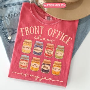May include: A watermelon-coloured t-shirt with the text "FRONT OFFICE chaos" and the phrase "is my jam". The shirt features illustrations of jars labelled "phones", "visitors", "multi task", "check-in", "schedule", "bells", "staff", and "buzz-in".