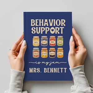 May include: A navy blue notebook with the words "BEHAVIOR SUPPORT" and "is my jam" printed in white. The notebook features illustrations of jars labeled with terms like "data," "charts," and "goals." The name "MRS. BENNETT" is also printed on the cover.