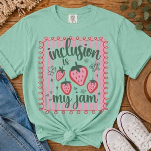 May include: A light mint green t-shirt featuring a central graphic with pink vertical stripes, a red scalloped border, and three pink strawberries. The text reads "inclusion is my jam" in dark green script, accompanied by small butterfly icons.