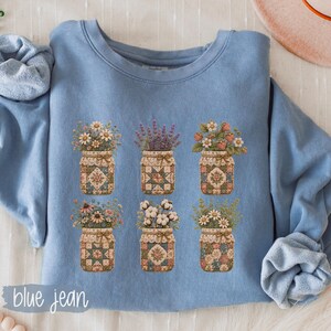 May include: A light blue crewneck sweatshirt featuring a graphic print of six mason jars arranged in two rows. Each jar contains different floral arrangements and is decorated with a unique, colorful quilt-like pattern and a small tied bow.