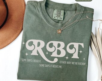 May include: Moss green t-shirt with the word "RGBT" in a retro font. The shirt also has the phrases "Some days I rock it. Some days it rocks me. Either way we're rockin'" printed on it. A gold necklace is draped across the shirt.