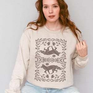 May include: A cream-colored sweatshirt with a graphic print of two stylized foxes in a floral frame. The foxes are brown and the floral frame is a light brown.