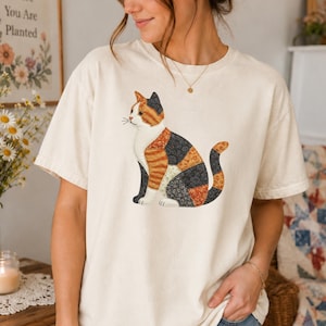 May include: A cream-coloured short-sleeve t-shirt featuring a graphic of a sitting cat. The cat is composed of various patchwork-style floral and striped patterns in shades of orange, black, white, and brown, creating a textured appearance.