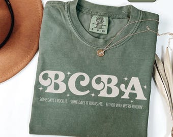 May include: Moss green t-shirt with the word "BCBA" in a retro font, with the text "SOME DAYS I ROCK IT. SOME DAYS IT ROCKS ME. EITHER WAY WE'RE ROCKIN'" below. A gold necklace with a pendant is on the shirt.