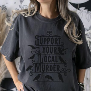 May include: A dark gray t-shirt with a graphic design featuring a black cage with the text "Support Your Local Murder" and several black crows.