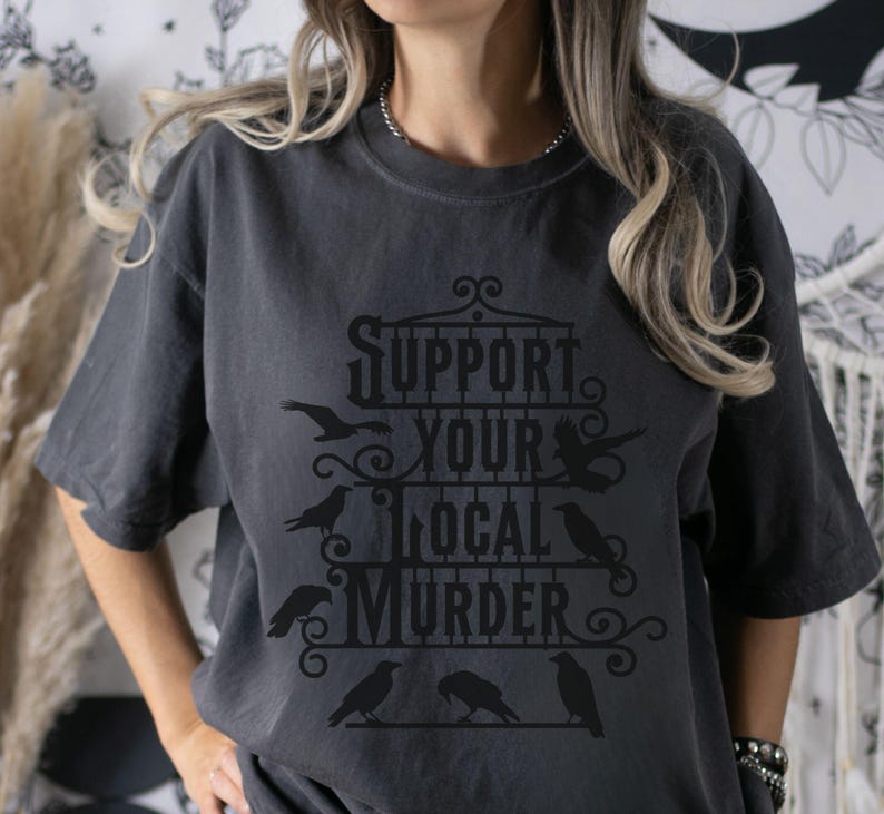 May include: A dark gray t-shirt with a graphic design featuring a black cage with the text "Support Your Local Murder" and several black crows.