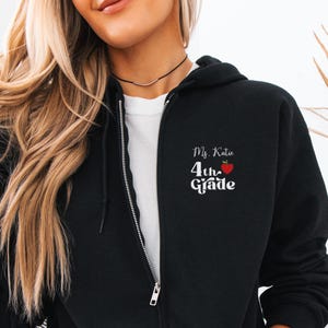Può includere: Felpa con cappuccio nera con zip con la scritta "Ms. Katie 4th Grade" e una grafica di mela rossa. La felpa ha un interno bianco e una cerniera argentata. La modella indossa una maglietta bianca sotto.