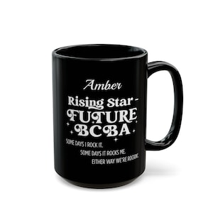 May include: Black ceramic mug with the text "Amber Rising Star FUTURE BCBA" in white. The mug also includes the phrases "SOME DAYS I ROCK IT," "SOME DAYS IT ROCKS ME," and "EITHER WAY WE'RE ROCKIN."