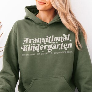 Può includere: Felpa con cappuccio verde oliva con la scritta "Transitional Kindergarten" in un font retrò. La felpa con cappuccio ha anche il testo "SOME DAYS I ROCK IT. SOME DAYS IT ROCKS ME. EITHER WAY WE'RE ROCKIN'" sotto. Ha una tasca frontale e un cappuccio.