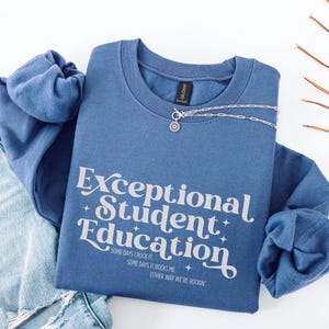 Può includere: Una felpa blu girocollo con la scritta "Exceptional Student Education" in bianco. La felpa riporta le frasi "Some days I rock it", "Some days it rocks me" e "Either way we're rockin'". Una collana d'argento con un ciondolo a forma di sole è sulla felpa.