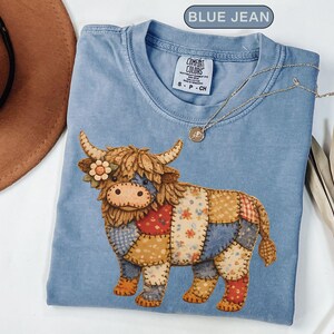 May include: A blue jean coloured t-shirt featuring a graphic of a shaggy highland cow with a patchwork body design. The cow has a small flower on its head and is rendered in various patterned fabric textures. The shirt has a Comfort Colors label.
