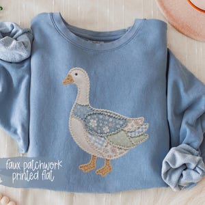 May include: A light blue crewneck sweatshirt featuring a central graphic of a goose with a faux patchwork design. The goose is composed of various floral and checkered patterns in shades of blue, white, and green, with yellow feet and beak.