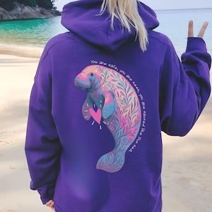 May include: A purple hoodie featuring a colorful illustration of a manatee with a heart and floral accents. The text "You are safe, you are seen, you are sacred like the sea" is printed on the back. The hoodie has a hood and long sleeves.