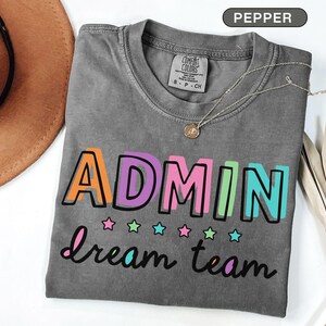 May include: A gray t-shirt with the words "ADMIN dream team" in colorful block letters and script. The shirt has a crew neck and short sleeves. A silver necklace with a pendant is on the shirt.