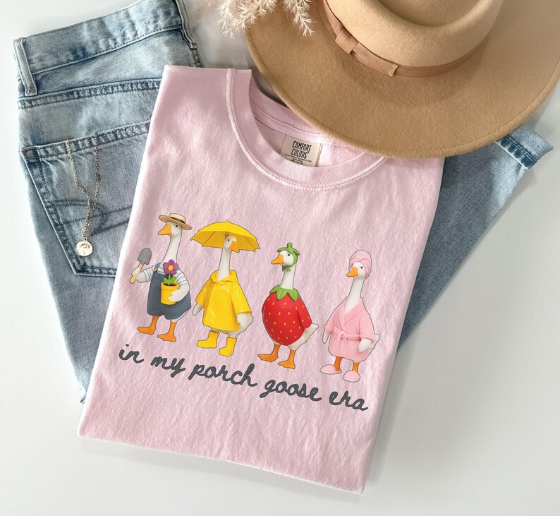 Pu&ograve; includere: Maglietta rosa chiaro con una grafica di quattro oche dei cartoni animati in vari abiti, tra cui un giardiniere, un'oca con un ombrello, un'oca fragola e un'oca in accappatoio. Il testo "in my porch goose era" &egrave; stampato sotto. La maglietta &egrave; abbinata a jeans blu e un cappello beige.