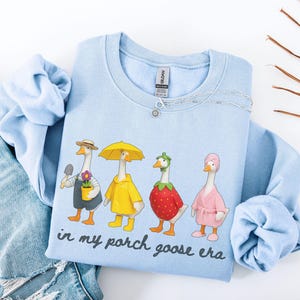 May include: A light blue sweatshirt featuring a whimsical illustration of four geese dressed in various outfits. One goose holds a trowel, another wears a yellow raincoat, a third is a strawberry, and the last wears a pink robe. The text "in my porch goose era" is printed below the geese.