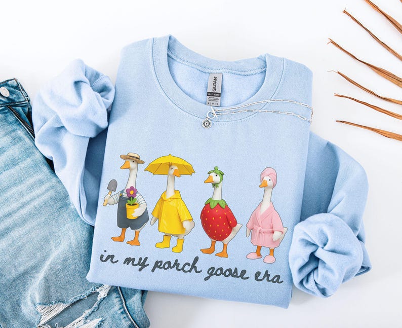May include: A light blue sweatshirt featuring a whimsical illustration of four geese dressed in various outfits. One goose holds a trowel, another wears a yellow raincoat, a third is a strawberry, and the last wears a pink robe. The text "in my porch goose era" is printed below the geese.