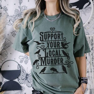 May include: A green t-shirt with a black graphic that says "Support Your Local Murder" with a black crow design.