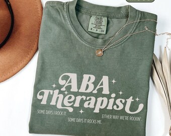May include: Moss green t-shirt with the text "ABA Therapist" in a retro font. The shirt also has the phrases "Some days I rock it", "Some days it rocks me", and "Either way we're rockin'". A gold necklace with a pendant is on the shirt.