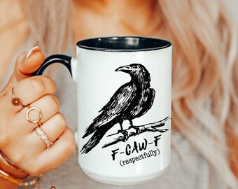 May include: White ceramic mug with a black rim and handle. The mug features a black illustration of a crow perched on a branch, with the text "F-CAW-F (respectfully)" below the bird. The mug is held by a person.