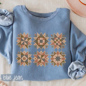 May include: A light blue crewneck sweatshirt featuring a graphic print of six distinct star-shaped quilt block patterns in muted tones of sage green, dusty rose, and cream. The text "blue jean" is printed in white in the bottom left corner.