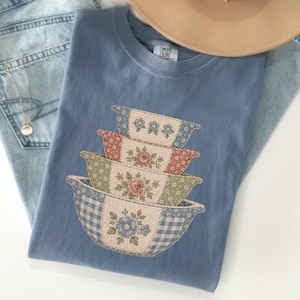 May include: A blue t-shirt featuring a graphic print of four stacked mixing bowls. The bowls are decorated with various floral patterns and a blue gingham check design. The shirt has a crew neckline and a Comfort Colors brand label at the neck.