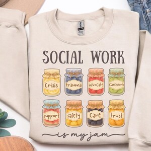 May include: A beige crewneck sweatshirt featuring the text SOCIAL WORK is my jam in dark brown. Below the text are eight illustrations of glass jars filled with colorful fruit preserves, each labeled with words including Crisis, trauma, advocate, casework, support, safety, care, and trust.