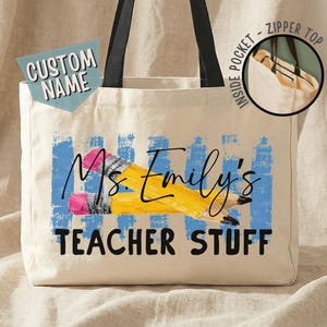 Può includere: Una borsa tote in tela beige con manici neri, caratterizzata da un disegno di due matite gialle su strisce verticali blu. Il testo recita Ms. Emily's TEACHER STUFF. Un inserto circolare mostra una tasca interna con chiusura a zip.