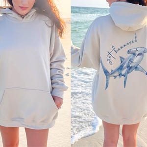 May include: Beige hoodie with a front pocket and a hood. The back of the hoodie features a blue watercolor hammerhead shark illustration with the text "get hammered" in a script font.
