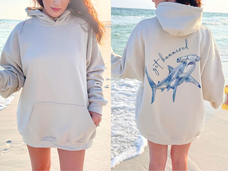 May include: Beige hoodie with a front pocket and a hood. The back of the hoodie features a blue watercolor hammerhead shark illustration with the text "get hammered" in a script font.