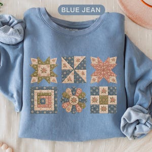 May include: A blue jean colored crewneck sweatshirt featuring a graphic print of six distinct quilt block patterns in muted tones of sage green, dusty rose, and navy blue. The sweatshirt has long sleeves with ribbed cuffs and a ribbed neckline.