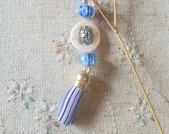 May include: A blue and white beaded necklace with a silver and white pendant and a purple tassel. The necklace is on a beige fabric background.