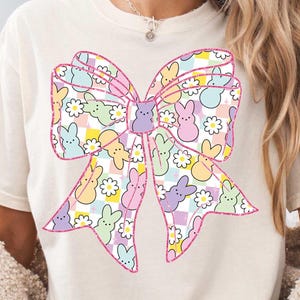 May include: A white t-shirt with a pink bow design. The bow is filled with a pattern of pastel-colored Peeps candy. The Peeps are arranged in a checkered pattern with white daisies.