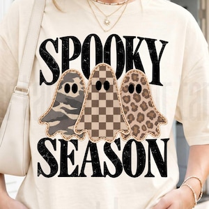 以下が含まれることがあります： クリーム色の半袖Tシャツ。黒い文字で「SPOOKY SEASON」と書かれ、3体のゴーストのイラストが描かれています。ゴーストはそれぞれ異なる柄で、迷彩柄、茶色の市松模様、ヒョウ柄が施されています。