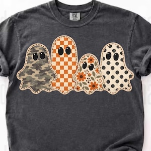 May include: A charcoal gray short-sleeve t-shirt featuring a graphic of four ghosts arranged in a row. The ghosts have distinct patterns: one with camouflage, one with an orange checkerboard, one with floral prints, and one with black polka dots.