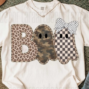以下が含まれることがあります： クリーム色の半袖Tシャツで、大きな文字でBOOと書かれています。Bはヒョウ柄、Oは迷彩柄の幽霊、もう一つのOはチェック柄の幽霊で、頭に白と黒の水玉模様のリボンをつけています。